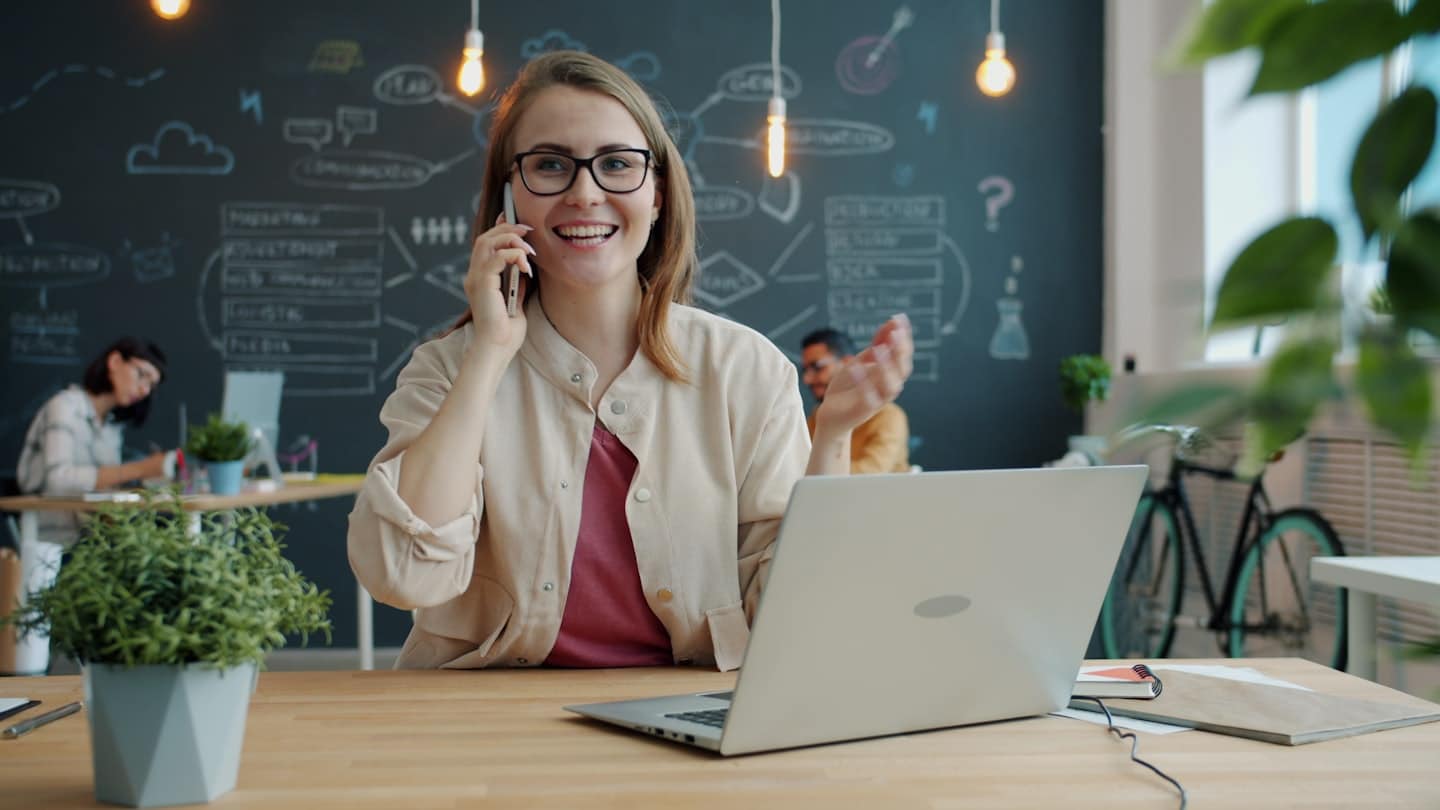 Gözlüklü bir kadın modern ofiste telefonla konuşuyor.